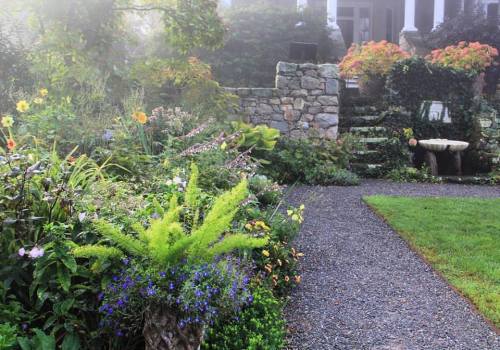 A lush garden with colorful flowers and greenery, featuring a stone path leading to a house in a misty atmosphere.