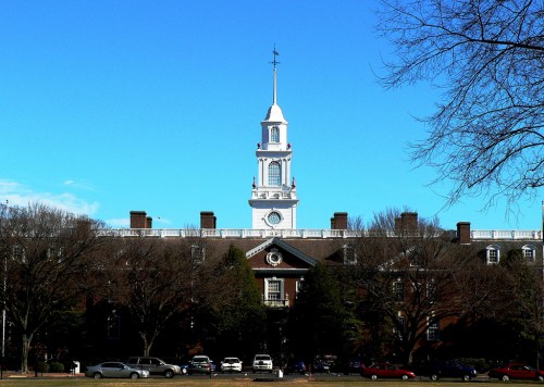 A historic building with a tall clock tower against a clear blue sky, surrounded by trees and parked cars.
