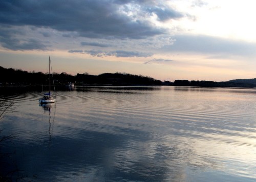 A calm lake at sunset with a sailboat and gentle ripples reflecting the sky and surrounding hills.