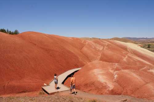 Two people walk along a wooden path through colorful, layered hills under a clear blue sky.