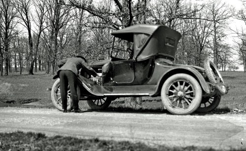 A vintage car parked on a dirt road, with a person inspecting the engine under the hood. Trees in the background.