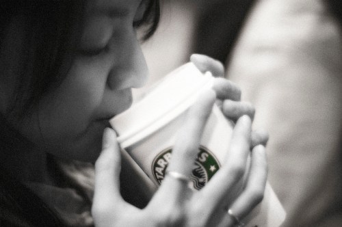 A close-up of a woman sipping from a Starbucks cup, her hands gently holding the cup. The image is in black and white.