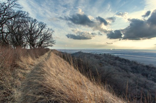 A scenic view of a grassy ridge overlooking a river valley, with trees and dramatic clouds in the sky.