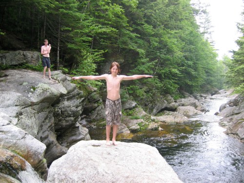 Two boys stand on rocks by a river, one with arms outstretched and the other in the background, surrounded by trees.
