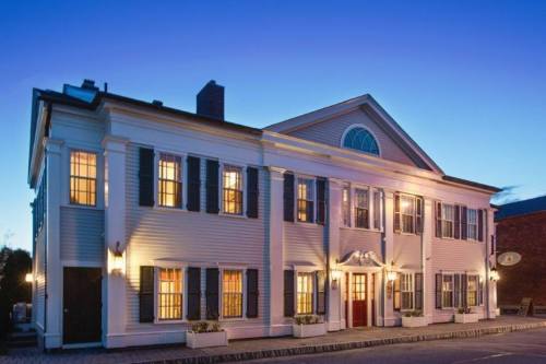 A charming two-story building with white siding, dark shutters, and warm lights glowing from the windows at dusk.