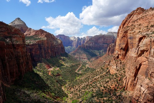 A panoramic view of Zion National Park, showcasing red rock formations and a winding valley under a blue sky with clouds.