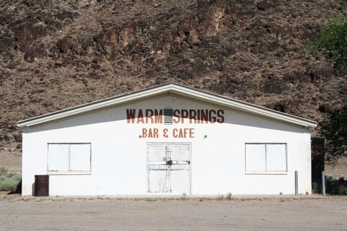A white building with "Warm Springs Bar & Cafe" painted on the front, set against a rocky hillside.