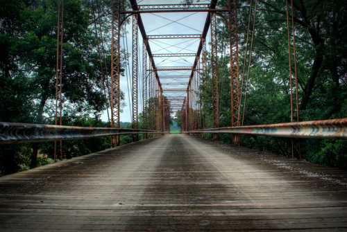 A weathered metal bridge stretches into the distance, surrounded by lush greenery under a cloudy sky.