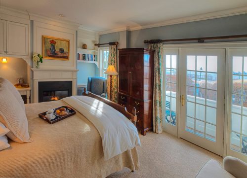Cozy bedroom with a fireplace, large windows, and a tray of breakfast on the bed, featuring soft decor and natural light.