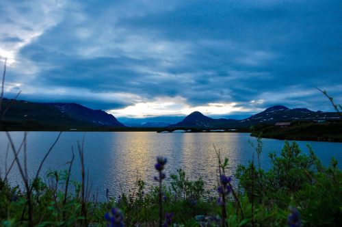 A serene lake reflects the cloudy sky, surrounded by mountains and lush greenery at dusk.