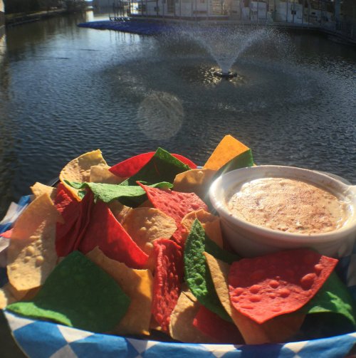 A plate of colorful tortilla chips with a bowl of dip, set against a scenic water fountain backdrop.