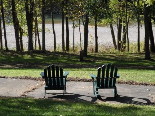 Two green Adirondack chairs sit on a patio, facing a serene river view surrounded by trees.