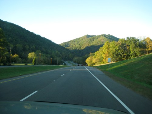 A scenic view of a winding road surrounded by green mountains and trees under a clear blue sky.