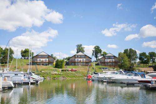 A serene marina with boats docked in the foreground and three wooden buildings on a grassy hill under a blue sky.