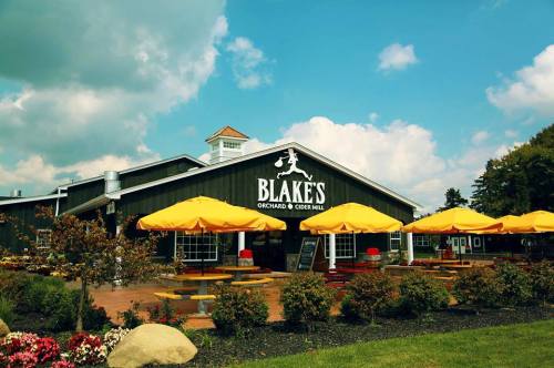 A large black barn-style building with yellow umbrellas outside, labeled "Blake's Orchard & Cider Mill," under a blue sky.