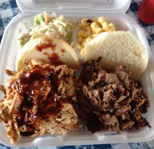 A takeout container with pulled pork, barbecue sauce, coleslaw, macaroni and cheese, and two sandwich buns.