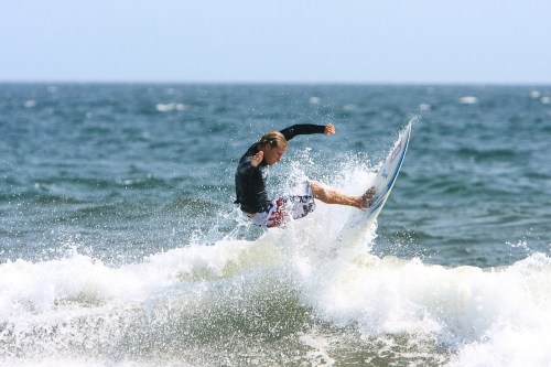 A surfer performs a trick on a wave, splashing water around in a sunny ocean setting.