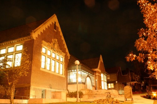 A brick building illuminated at night, featuring large windows and a decorative roof, surrounded by trees.
