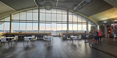 A spacious airport terminal with large windows, empty tables, and a few people gathered near the counter.