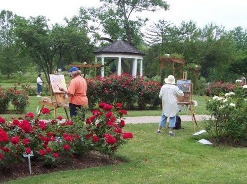 Two artists painting in a rose garden, surrounded by vibrant red flowers and a gazebo in the background.