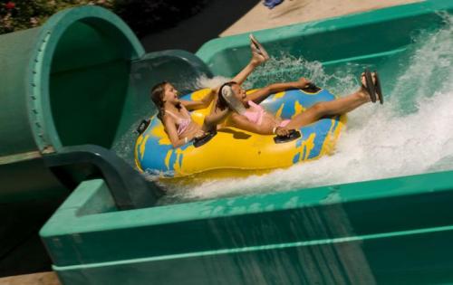 Two girls slide down a water slide on a yellow inflatable tube, splashing into the pool below.