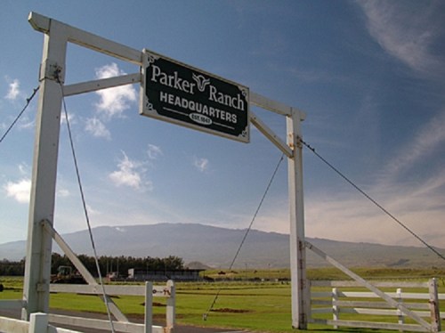 Sign for Parker Ranch Headquarters against a backdrop of blue skies and distant mountains. Green grass in the foreground.
