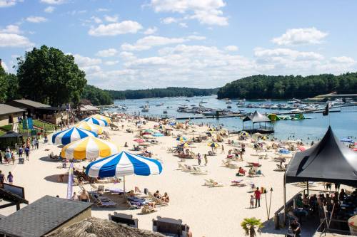 A sunny beach scene with colorful umbrellas, people relaxing, and boats on the water under a blue sky with clouds.