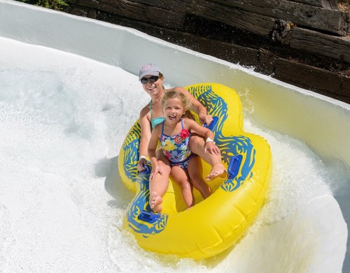 A woman and a girl joyfully ride a yellow inner tube down a water slide, splashing through the water.