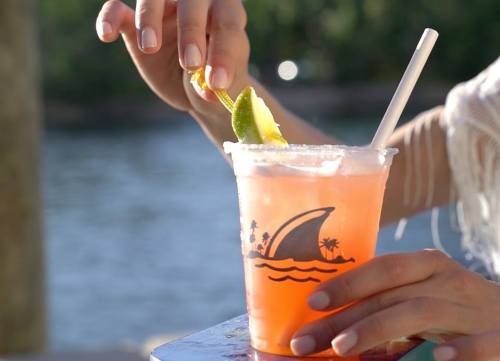 A hand garnishes a colorful drink with a lime slice, set against a blurred outdoor background.