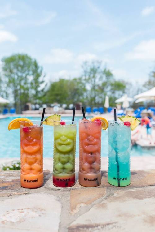 Four colorful cocktails with fruit garnishes sit on a stone ledge by a pool, with lounge chairs in the background.