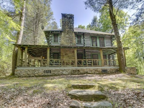 A rustic log cabin surrounded by trees, featuring a stone chimney and a spacious porch.