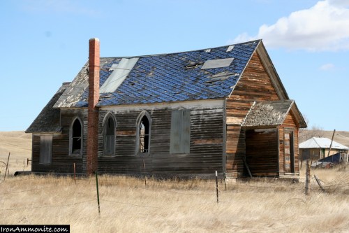 Abandoned wooden building with a blue roof, broken windows, and a brick chimney, set in a grassy landscape.