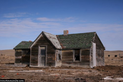 Abandoned wooden house with a green roof, broken windows, and a barren landscape under a blue sky.