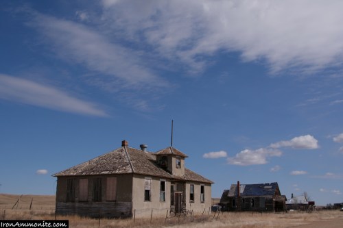 Abandoned houses under a blue sky with scattered clouds, set in a rural landscape.