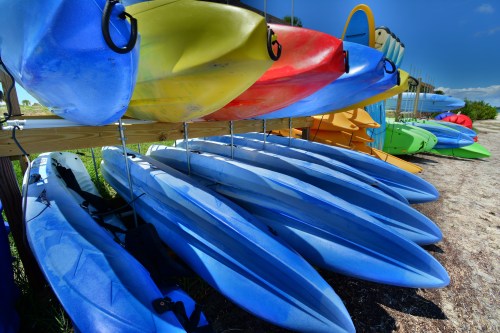 Colorful kayaks stacked on a rack, with blue, yellow, red, and green boats against a clear blue sky.