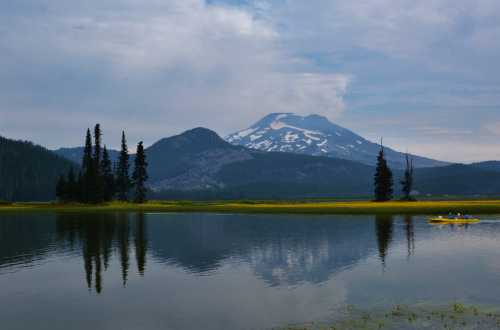 A serene lake reflects mountains and trees under a cloudy sky, with a kayak visible on the water.