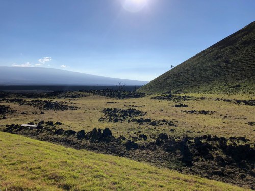 A scenic landscape featuring rolling green hills, rocky terrain, and a bright blue sky with the sun shining.