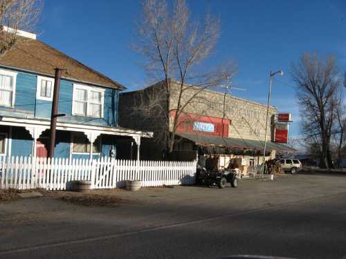 A blue house with a white picket fence next to a brick building, with trees and a clear blue sky in the background.