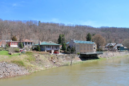 A scenic view of riverside buildings with trees and hills in the background under a clear blue sky.