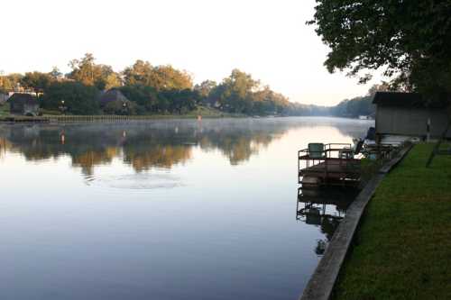 A calm river at dawn, with mist rising and chairs on a wooden dock beside lush greenery.