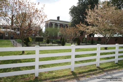 A white fence in front of a park with blooming trees and a historic building in the background under a cloudy sky.