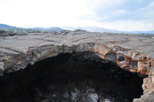 A rocky volcanic landscape with a large, open crevice revealing dark, jagged rock formations beneath.