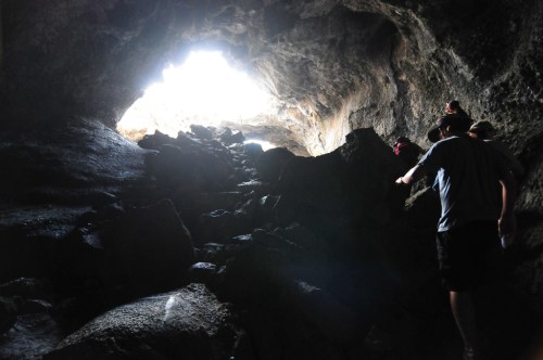 A person climbs rocky terrain inside a dark cave, with bright light shining from the cave's opening above.