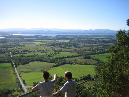 Two people stand on a lookout, gazing at a vast landscape of fields, mountains, and a lake under a clear blue sky.