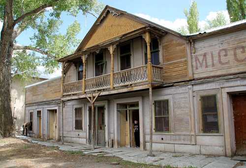 A weathered wooden building with a balcony, featuring multiple doors and windows, set in a rustic outdoor environment.