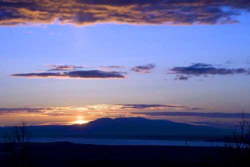 A serene sunset over mountains, with vibrant colors reflecting on the water below and clouds scattered in the sky.