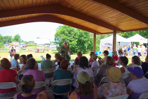 A group of people seated under a wooden pavilion, listening to a speaker in a garden setting.