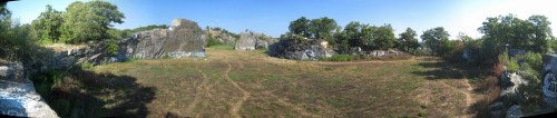 Panoramic view of a grassy area surrounded by large rocks and trees under a clear blue sky.