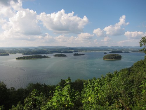 A scenic view of a lake with several islands, surrounded by green hills and a blue sky with fluffy clouds.