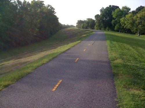 A paved path winds through a grassy area, bordered by trees on one side and a gentle slope on the other.